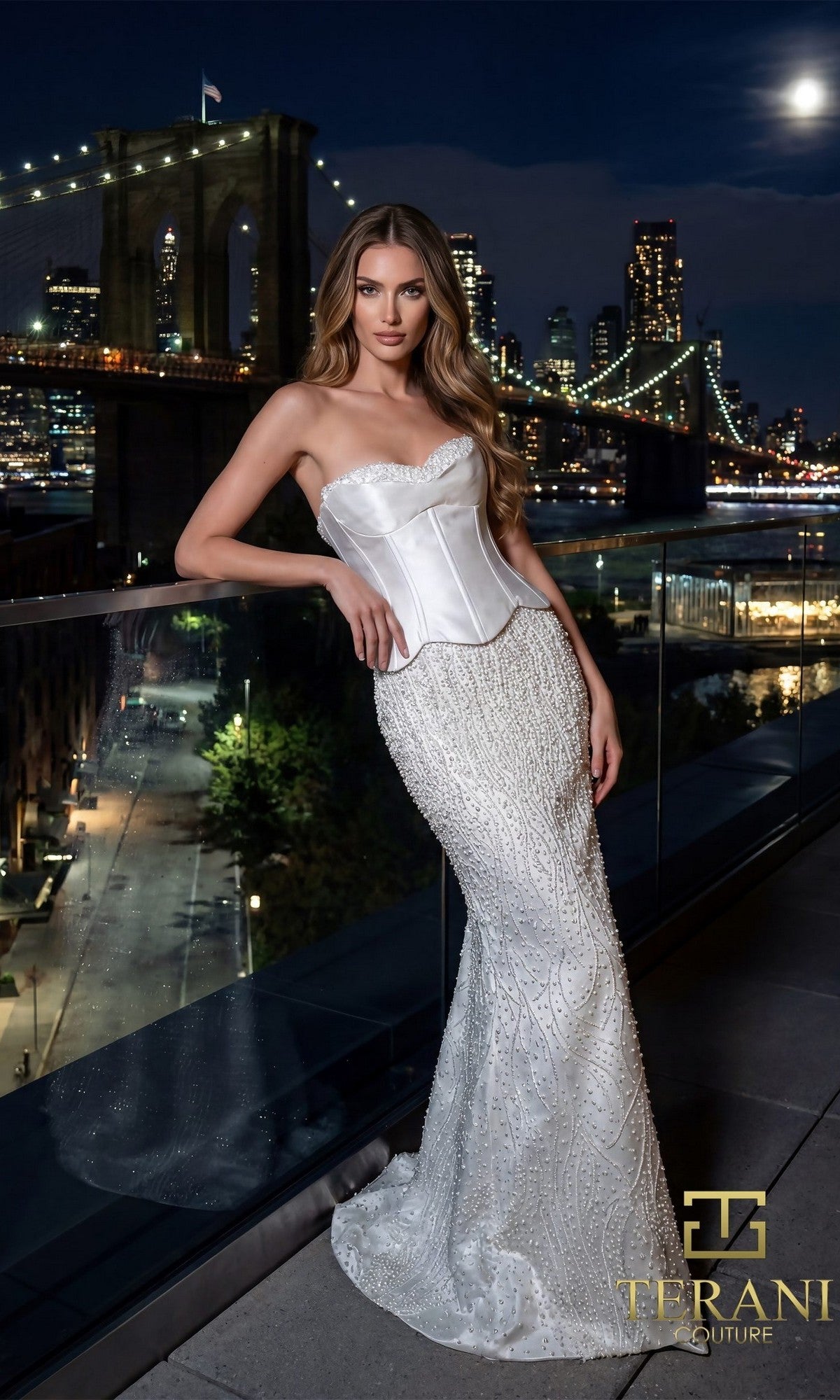 A woman in an ivory Long Formal Dress (Terani 261E6238) with pearl embellishments poses on a balcony at night, the sparkling city skyline and Brooklyn Bridge forming a stunning backdrop.